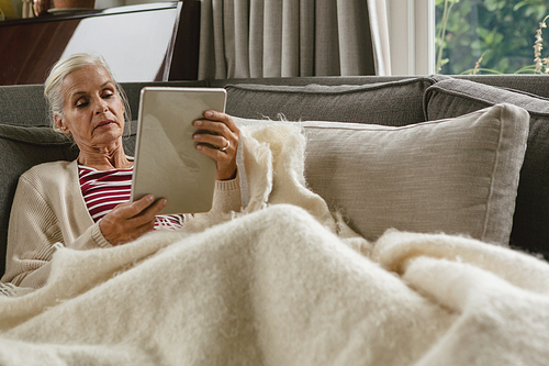 Front view of active senior Caucasian woman relaxing on sofa and using digital tablet in living room at comfortable home
