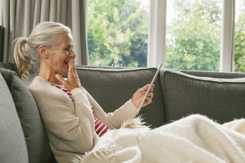 Side view of happy active senior Caucasian woman relaxing on sofa and using digital tablet in living room at comfortable home