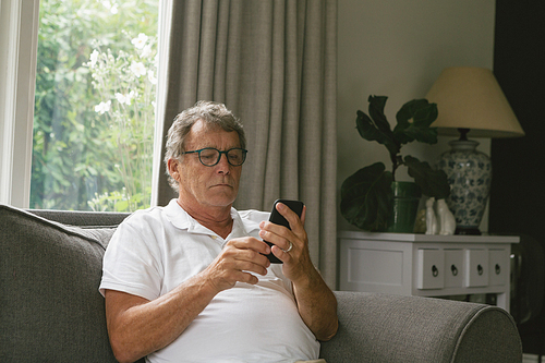 Front view of active senior Caucasian man sitting on sofa and using mobile phone in living room at comfortable home