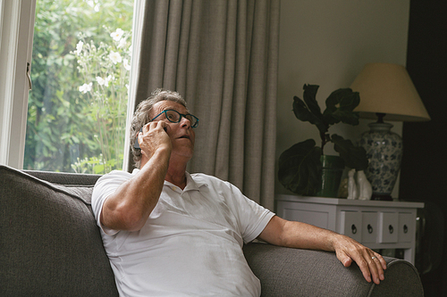 Front view of active senior Caucasian man sitting on sofa and talking on mobile phone in living room at comfortable home