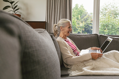 Side view of happy active senior Caucasian woman relaxing on sofa and using laptop in living room at comfortable home