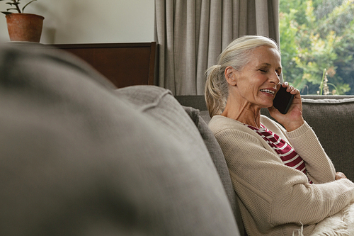 Side view of active senior Caucasian woman sitting on sofa and talking on mobile phone in living room at comfortable home