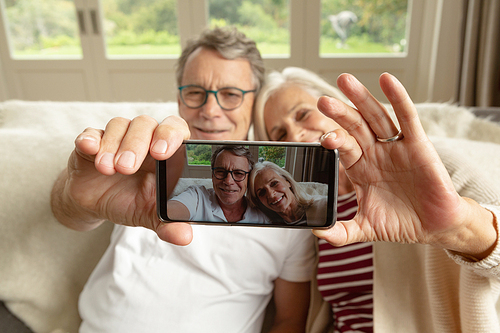 Front view of happy active senior Caucasian couple sitting on sofa and taking selfie in a comfortable home