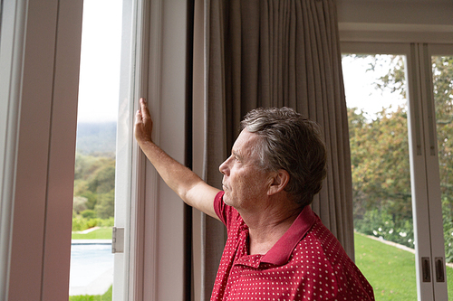 Side view of thoughtful active senior Caucasian man looking through window in a comfortable home