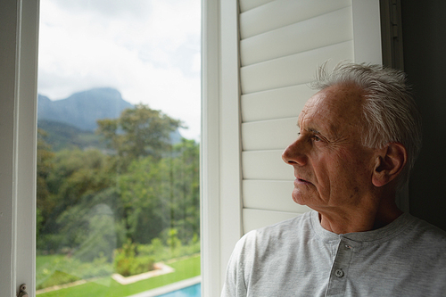 Front view of thoughtful active senior Caucasian man looking through window in a comfortable home