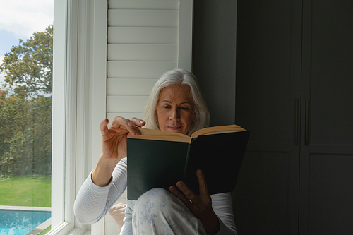 Front view of active senior Caucasian woman reading a book on window seat at home