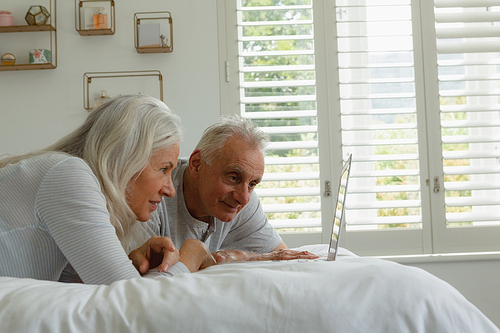 Side view of active senior Caucasian couple using laptop while lying on bed in bedroom at home