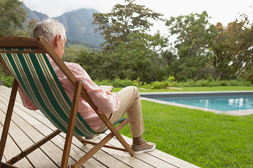 Rear view of active senior Caucasian man reading a book while relaxing on sun lounger in the backyard of home