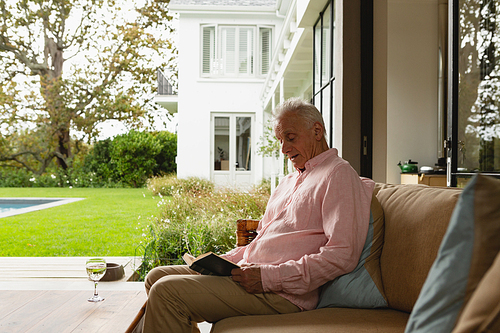 Side view of active senior Caucasian man reading a book while relaxing on sofa in the porch art home
