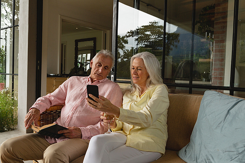 Front view of active senior Caucasian couple using mobile phone and reading a book in the porch at home