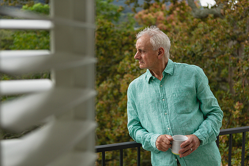 Side view of active senior Caucasian man standing with coffee mug on the balcony at home