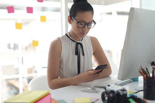 Front view of beautiful Asian female graphic designer using mobile phone at desk in a modern office