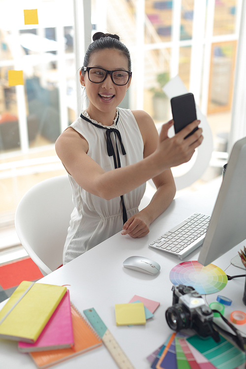 Front view of beautiful Asian female graphic designer taking selfie with mobile phone at desk in a modern office