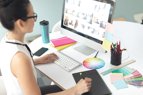 Rear view of attentive Asian female graphic designer using graphic tablet at desk in a modern office