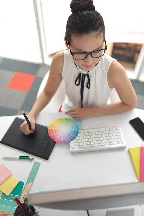 High angle view of Asian female graphic designer using graphic tablet at desk in a modern office