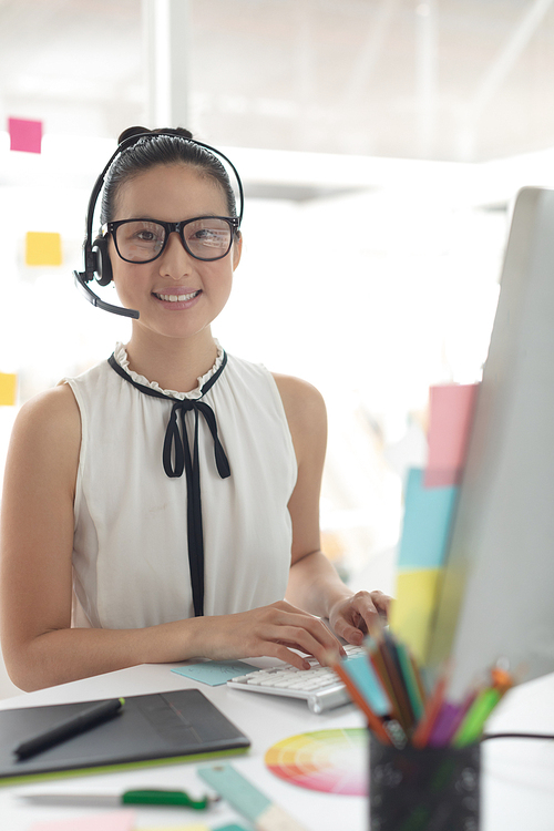 Front view of happy Asian female graphic designer in headset  while working at desk in a modern office