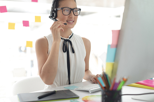 Front view of happy Asian female graphic designer talking on headset at desk in a modern office