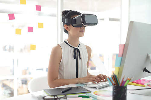 Side view of Asian female graphic designer using virtual reality headset while working on computer at desk in a modern office