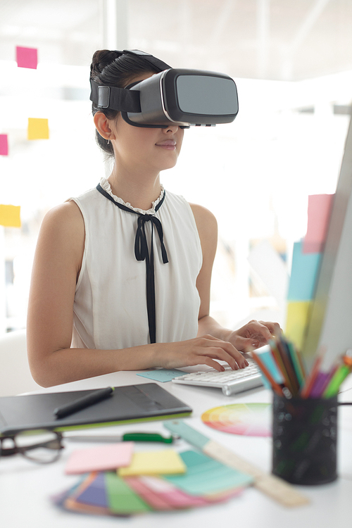 Side view of Asian female graphic designer using virtual reality headset while working on computer at desk in a modern office