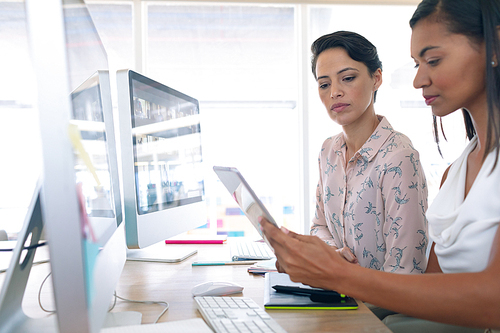 Side view of diverse female graphic designers discussing on digital tablet at desk in a modern office