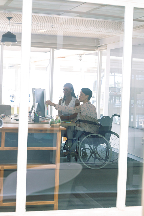 side view of diverse female graphic designers discussing on computer at desk in a modern office. . mixed-race female designer is sitting in wheelchair.