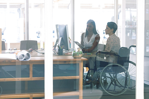 side view of mixed race female graphic designers working together at desk in a modern office. . mixed-race female designer is sitting in wheelchair.