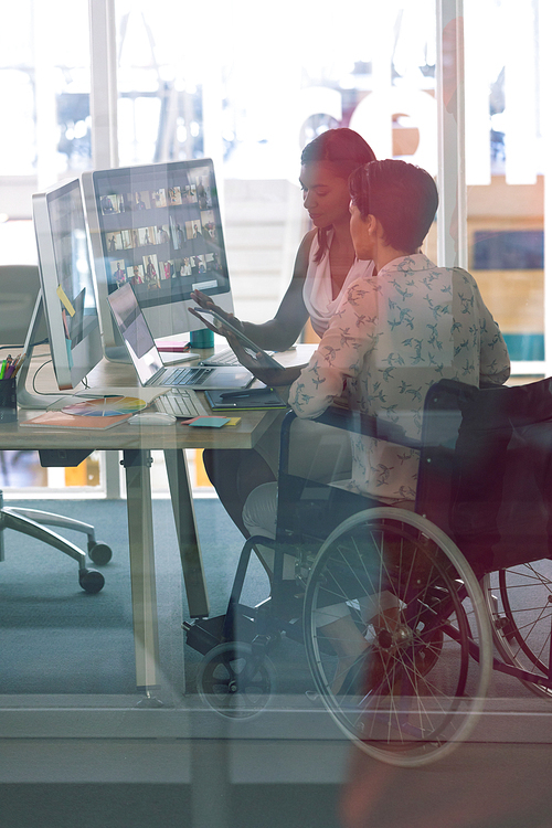 side view of diverse female graphic designers working together at desk in a modern office.. mixed-race female designer is sitting in wheelchair.