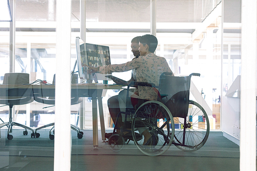 side view of diverse female graphic designers discussing on computer at desk in a modern office. . mixed-race female designer is sitting in wheelchair.