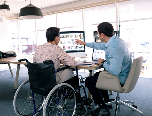 Rear view of diverse graphic designers discussing over computer at desk in a modern office. Disabled mixed-race female designer is sitting in wheelchair.