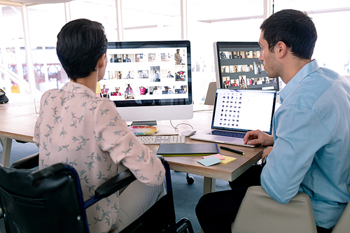 Rear view of diverse graphic designers working together at desk in a modern office. Disabled mixed-race female designer is sitting in wheelchair.