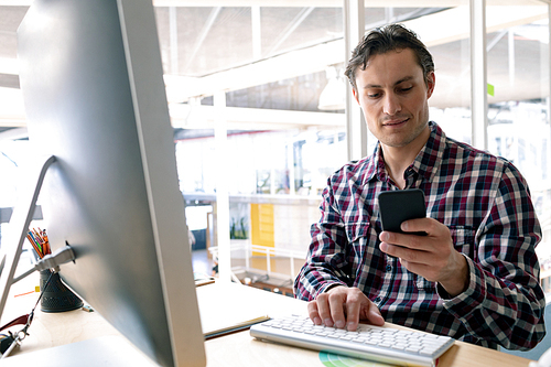 Front view of Caucasian male graphic designer using mobile phone while working on computer at desk in a modern office