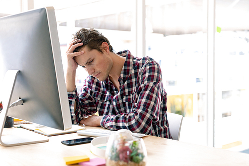 Side view of tensed Caucasian male graphic designer with hand on head sitting at desk in a modern office