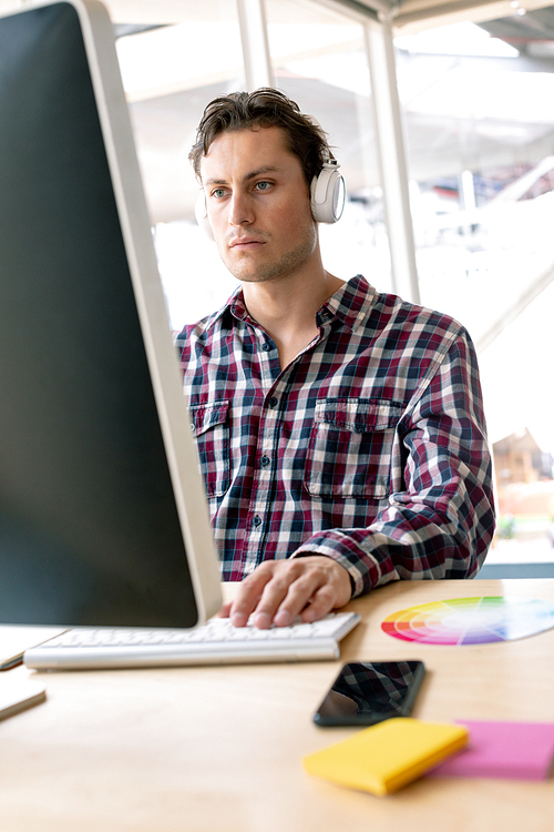 Front view of Caucasian male graphic designer listening music on headphone while working on computer at desk in a modern office