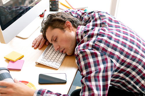 High view of tired Caucasian male graphic designer sleeping on desk in a modern office