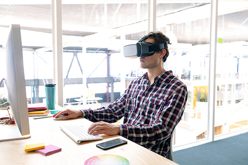 Side view of Caucasian male graphic designer using virtual reality headset while working on computer at desk in office