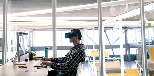 Side view of Caucasian male graphic designer using virtual reality headset while working on computer at desk in office