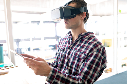 Side view of Caucasian male graphic designer using virtual reality headset while working on digital tablet at desk in office