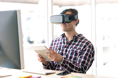 Side view of Caucasian male graphic designer using virtual reality headset while working on digital tablet at desk in office