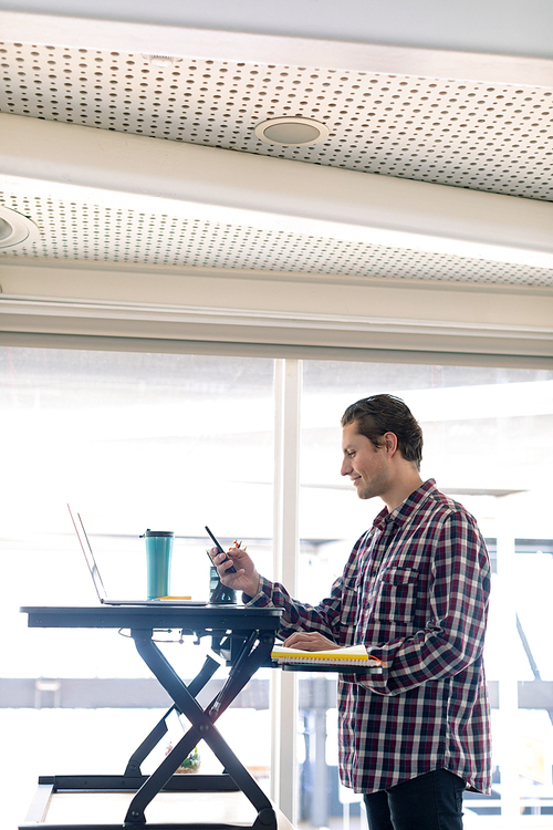 Side view of Caucasian male graphic designer using mobile phone while working on laptop at desk in office