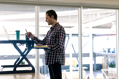 Side view of Caucasian male graphic designer using mobile phone while working on laptop at desk in office