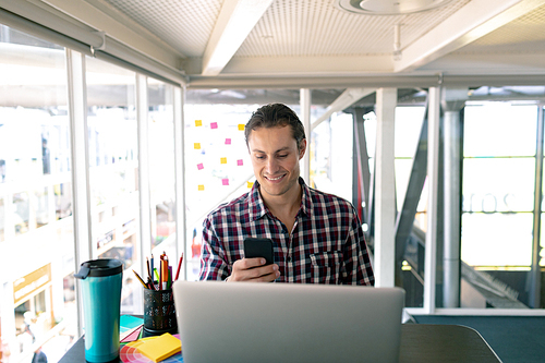 Front view of Caucasian male graphic designer using mobile phone while working on laptop at desk in office