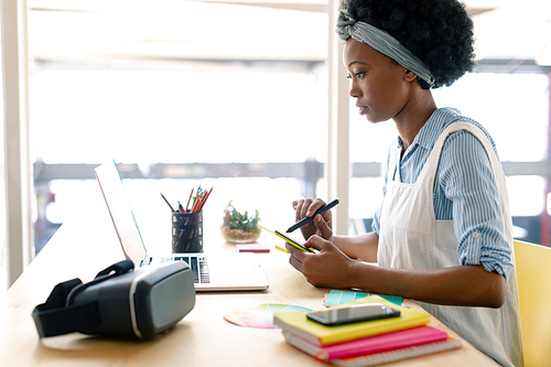 Side view of African american female graphic designer using graphics tablet while looking at laptop at desk in office