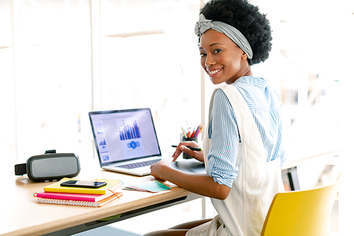 Front view of African american female graphic designer using graphic tablet while looking at laptop at desk in office