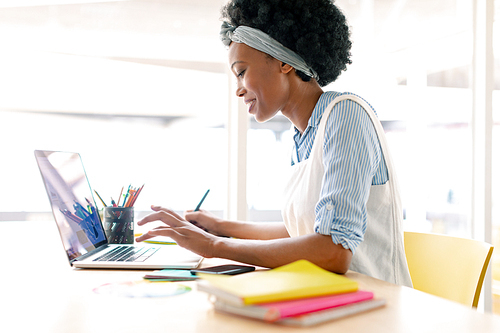 Side view of African american female graphic designer using graphic tablet and laptop at desk in office