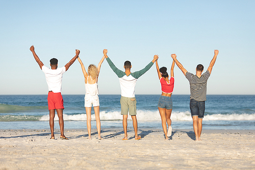 Rear view of diverse friends standing together with hands raised on the beach