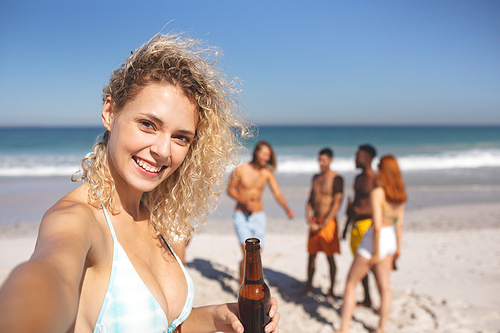 Front view of Caucasian girl taking selfie while group of young diverse friends having fun together on the beach