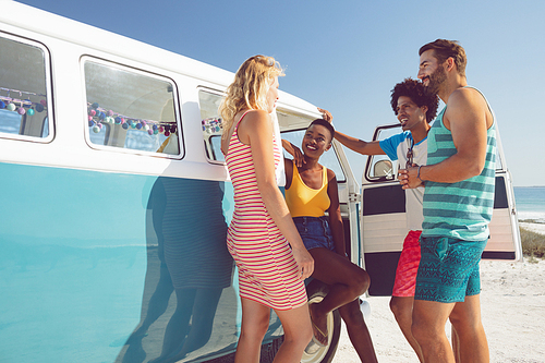 Front view of happy diverse group of friends talking with each other near camper van at beach in the sunshine