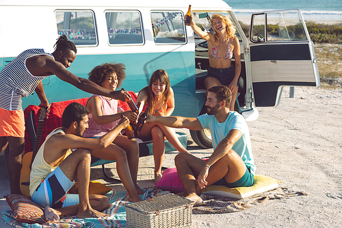 Front view of happy group of diverse friends toasting beer bottle near camper van at beach