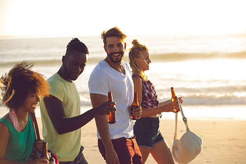 Side view of happy group of diverse friends holding beer bottles and walking together on the beach