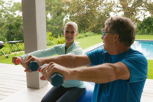 Front view of active senior Caucasian couple exercising with dumbbell in porch at home